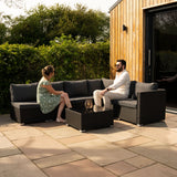 Modulares 7-teiliges Rattan-Möbelset in Schwarz mit grauen Kissen, ideal für den Außenbereich, umgeben von grüner Natur und einem modernen Holzhaus