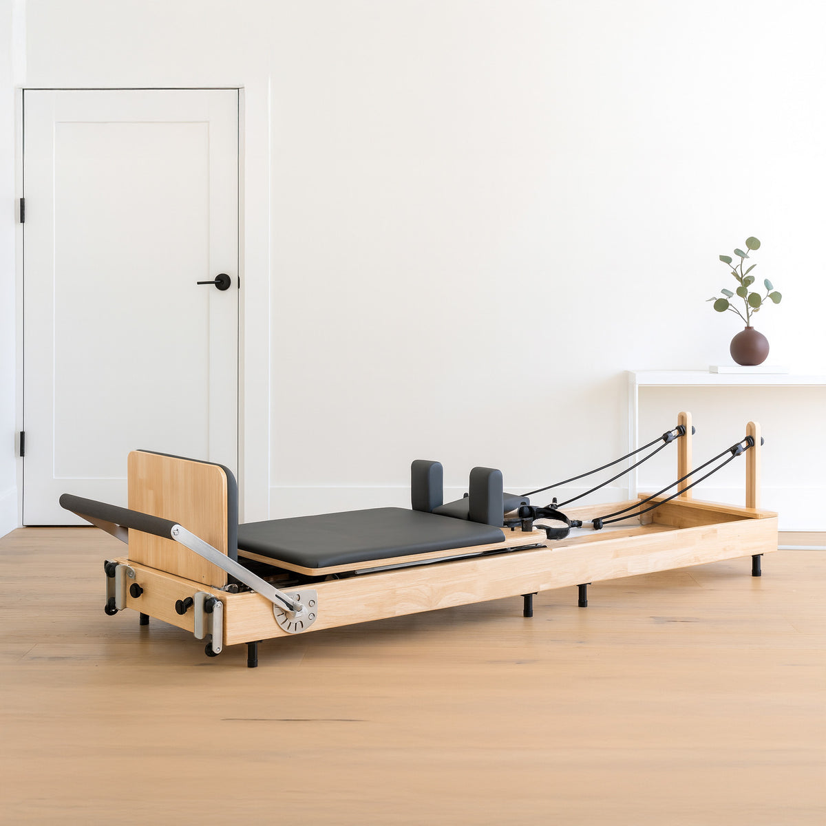 Pilates Reformer Maschine aus Eiche mit schwarzem Polster, verstellbaren Griffen und elastischen Widerstandsbändern in einem hellen Raum