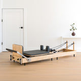 Pilates Reformer Maschine aus Eiche mit schwarzem Polster, verstellbaren Griffen und elastischen Widerstandsbändern in einem hellen Raum