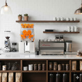 Kommerzielle Orangensaftpresse aus Edelstahl mit orangefarbenen Saftbehältern, platziert auf einer modernen, hellen Theke mit Kaffeegeräten und Regalen