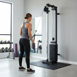 Wandmontierte Kabelzugmaschine in modernem Fitnessstudio, schwarze Gewichte, graue Wand, Sportlerin in grauem Top und schwarzen Leggings beim Training