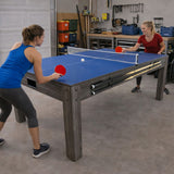 Zwei Frauen spielen Tischtennis an einem grauen 7-in-1 Spieltisch mit Stauraum, blauer Tischplatte und robusten Holzbeinen in einer Werkstattumgebung