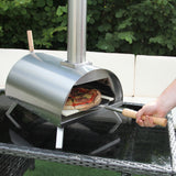 Der anthrazitgraue Tisch-Pizzaofen aus Edelstahl mit abgerundeter Form und Holzgriffen, auf einem eleganten Aluminium-Ecksofa im Hintergrund