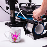 Eine weiße Sublimationstasse mit pinkfarbener Lilienblüte steht neben einer Druckmaschine, die für die Tassenbedruckung vorbereitet wird