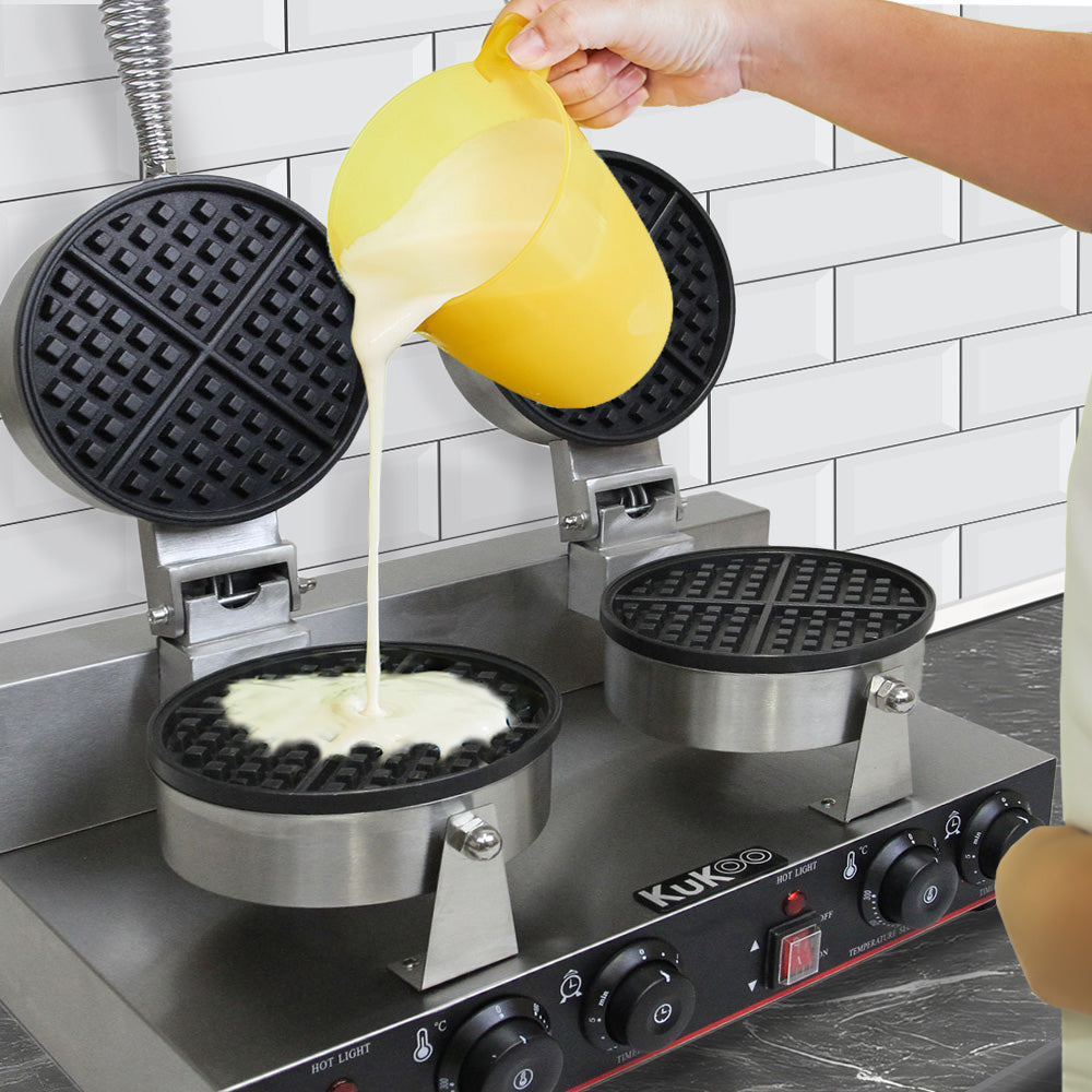 Zwei runde Waffeleisen aus Edelstahl mit schwarzem, rutschfestem Gitter, während Teig aus einem gelben Messbecher hineingegossen wird
