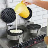Zwei runde Waffeleisen aus Edelstahl mit schwarzem, rutschfestem Gitter, während Teig aus einem gelben Messbecher hineingegossen wird