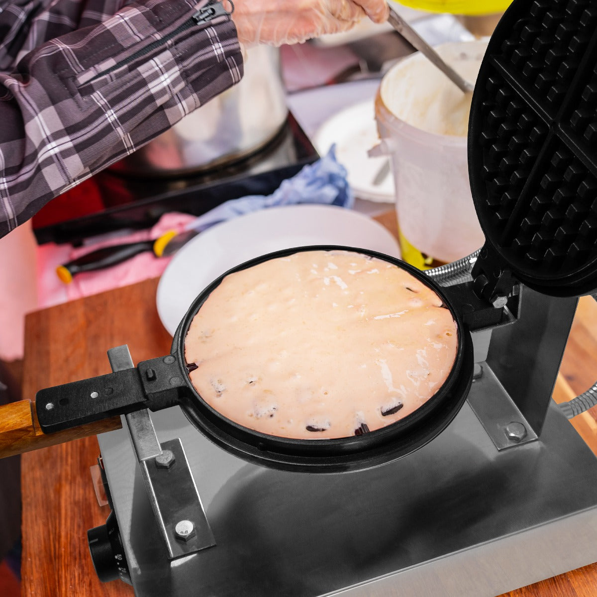 Das KuKoo Profi Waffeleisen mit einer glänzenden, schwarzen Oberfläche und einem Holzgriff, während frischer Waffelteig gleichmäßig backt