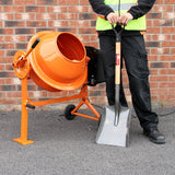Zementmischer in leuchtendem Orange mit Handbetonrüttler, 2 Meter Schaft, auf grauem Untergrund, neben einer Person in Arbeitskleidung mit Schaufel