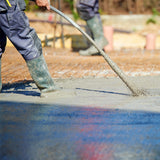 Arbeiter in Gummistiefeln, der mit einem Handbetonrüttler frischen Beton auf einer Baustelle bearbeitet, um eine glatte Oberfläche zu erzielen