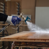 Maxblast Airless Farbsprühgerät in Aktion, sprüht gleichmäßigen Farbnebel auf eine Holzoberfläche, ergonomischer Griff, blaue Sprühdüse, präzise Anwendung