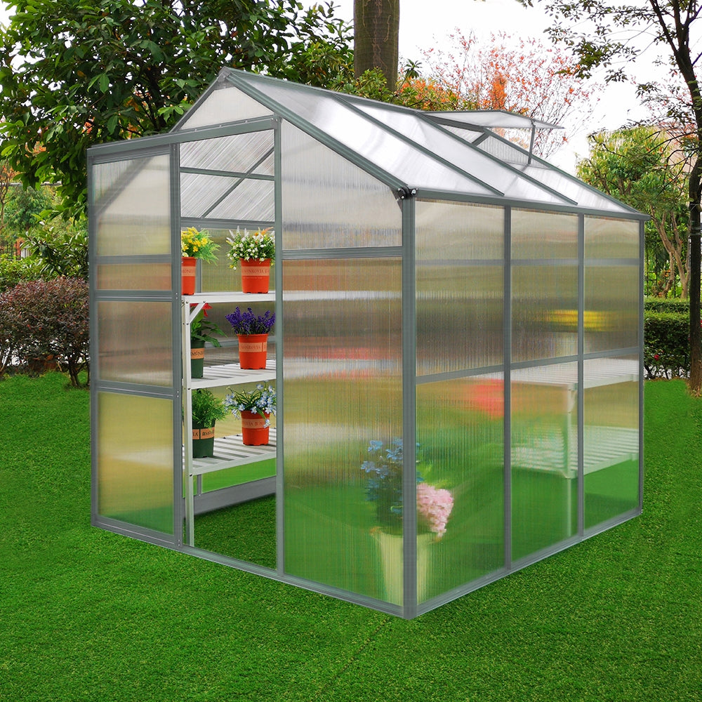 Gewächshaus 1.8m x 1.8m ohne Boden aus transparentem Polycarbonat, mit stabilen Metallrahmen und Regalen für Pflanzen, umgeben von grünem Gras und Bäumen
