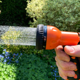 Gartenbewässerung mit einem orangefarbenen Sprühkopf, der Wasser in feinen Strahlen versprüht, umgeben von grünen Pflanzen und blühenden Blumen