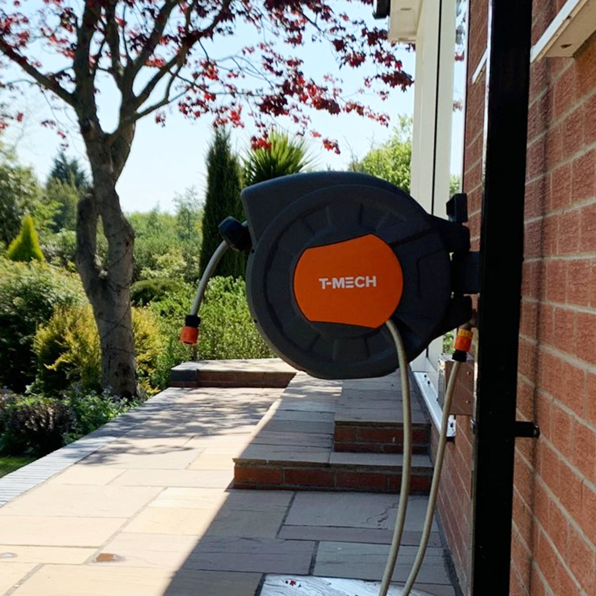 Schwarze Schlauchtrommel mit orangefarbenem T-MECH-Logo, montiert an einer Wand, mit einem 20m langen Gartenschlauch, umgeben von grünem Garten