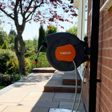 Schwarze Schlauchtrommel mit orangefarbenem T-MECH-Logo, montiert an einer Wand, mit einem 20m langen Gartenschlauch, umgeben von grünem Garten