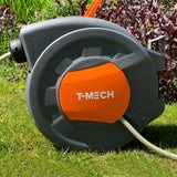 Gartenschlauch auf einer grauen Schlauchtrommel mit orangefarbenem Detail, platziert auf grünem Gras vor blühenden Pflanzen