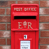 Roter Briefkasten mit Bodenständer, glänzender Oberfläche, goldene Buchstaben und Krone, vor einer Backsteinmauer, mit Sammelzeiten-Schild