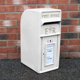 Weißer Briefkasten mit Bodenständer, aus robustem Material, mit königlichem Emblem und klar lesbarem Abholzeit-Schild, vor einer Ziegelmauer platziert
