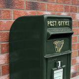 Grüner Briefkasten im irischen Stil mit goldener Harfe und der Aufschrift "POST OFFICE", vor einer roten Ziegelwand platziert