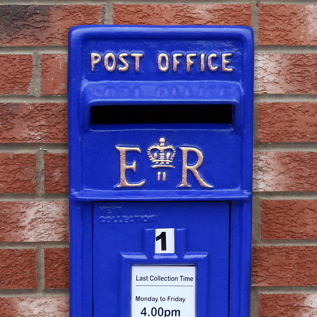 Blauer Briefkasten mit Bodenständer, verziert mit goldenen Buchstaben und einer Krone, vor einer roten Ziegelmauer platziert