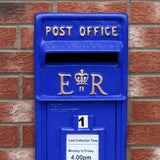 Blauer Briefkasten mit Bodenständer, verziert mit goldenen Buchstaben und einer Krone, vor einer roten Ziegelmauer platziert