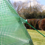 Grünes Netzmaterial mit quadratischem Muster, das einen Polytunnel sichert, vor unscharfem Hintergrund von Bäumen und Wiese