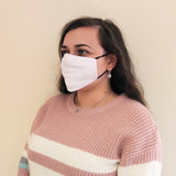 Weiße, wiederverwendbare Gesichtsmaske, getragen von einer Frau mit langen, braunen Haaren, in einem hellrosa, gestreiften Pullover, vor neutralem Hintergrund
