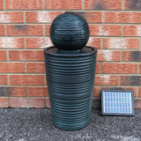 Schwarzer Kugelbrunnen aus Keramik mit strukturierten Rillen, der Wasser sprudelt, vor einer roten Ziegelwand und einem Solarpanel platziert