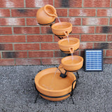 Terracotta Solar Kaskadenbrunnen mit fünf Töpfen, die Wasser in eine große Schale leiten, umgeben von grauem Kies und einer Solarpanel neben der Wand