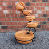 Terracotta Solar Kaskadenbrunnen mit fünf Töpfen, die Wasser in eine große Schale leiten, umgeben von grauem Kies und einer Ziegelmauer