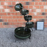Solar Kaskadenbrunnen in Grün mit fünf Töpfen, die Wasser in eine große Schale leiten, umgeben von grauem Kies und einer Solarpanel neben einer Ziegelwand