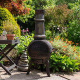Gusseisen Terrassenofen mit rustikaler Oberfläche, runder Form und hohem Schornstein, umgeben von bunten Blumen und grünem Garten