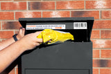Schwarzer Paketkasten mit geöffnetem Deckel, in dem ein gelbes Paket eingelegt wird, vor einer roten Ziegelwand platziert