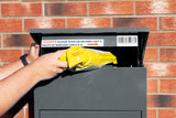 Grau Paketkasten mit geöffneter Klappe, in der eine gelbe Lieferung eingelegt wird, vor einer roten Ziegelwand platziert