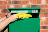 Grüner Paketkasten mit geöffneter Klappe, in der eine gelbe Versandtasche eingelegt wird, vor einer roten Ziegelwand platziert