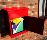 Roter Paketkasten aus Metall mit glänzender Oberfläche, geöffnet und zeigt ein gelbes Paket im Inneren, vor einer Backsteinwand platziert