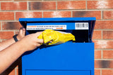 Blauer Paketkasten mit geöffneter Klappe, in den eine gelbe Versandtasche eingelegt wird, vor einer Backsteinmauer platziert