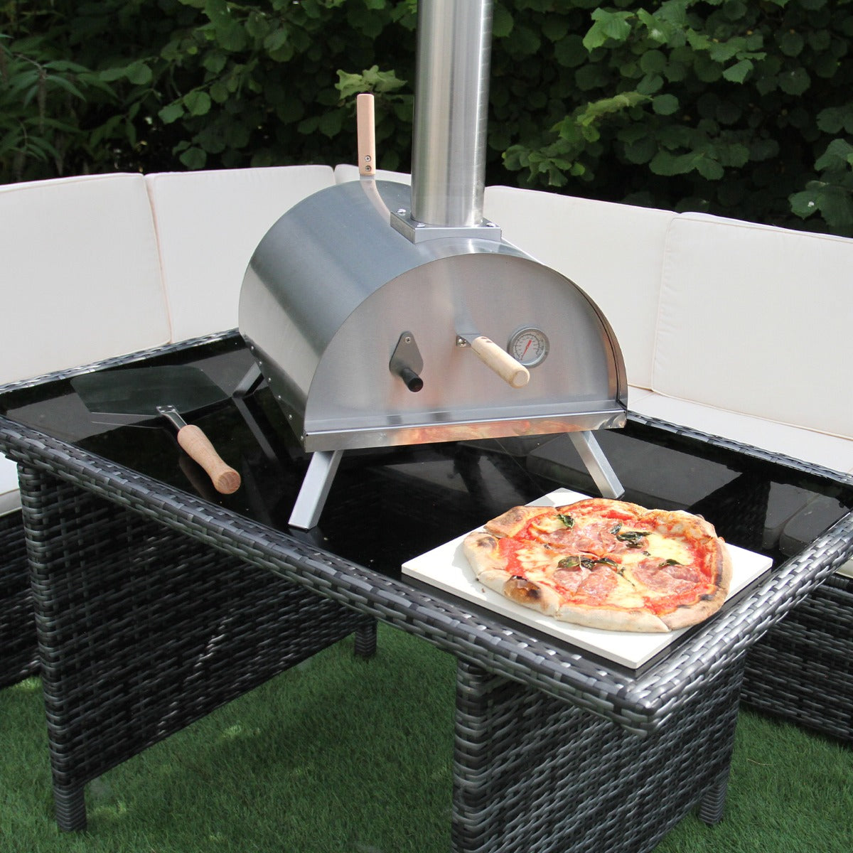 Eleganter Tisch-Pizzaofen aus Edelstahl mit Holzgriffen, auf einem Tisch platziert, neben einer frisch gebackenen Pizza auf einem weißen Teller