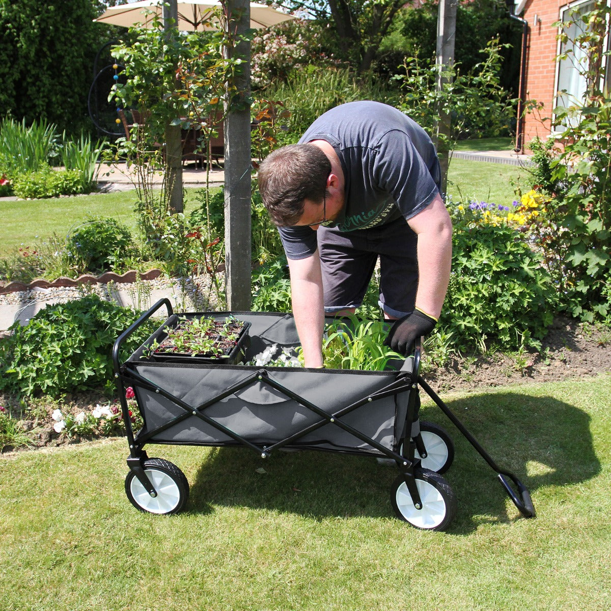 Faltbarer Gartenwagen in Grau mit robustem Stoff, großen Rädern und einem Benutzer, der Pflanzen im Garten transportiert, umgeben von grünen Pflanzen
