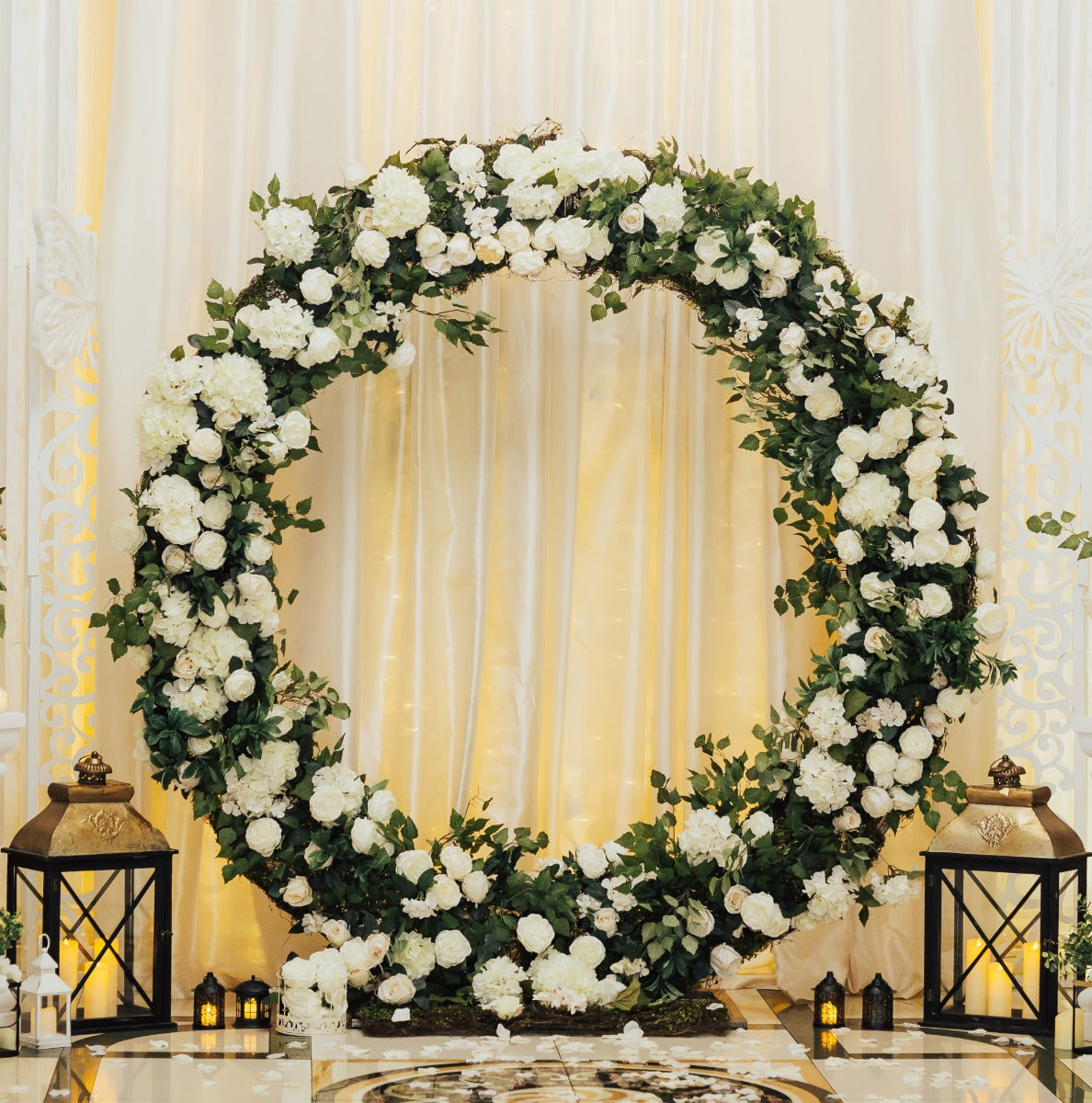 Eleganter Hochzeit Ballonbogen in Weiß, umgeben von frischen weißen Blumen und grünen Blättern, dekoriert mit stilvollen Laternen und sanftem Licht