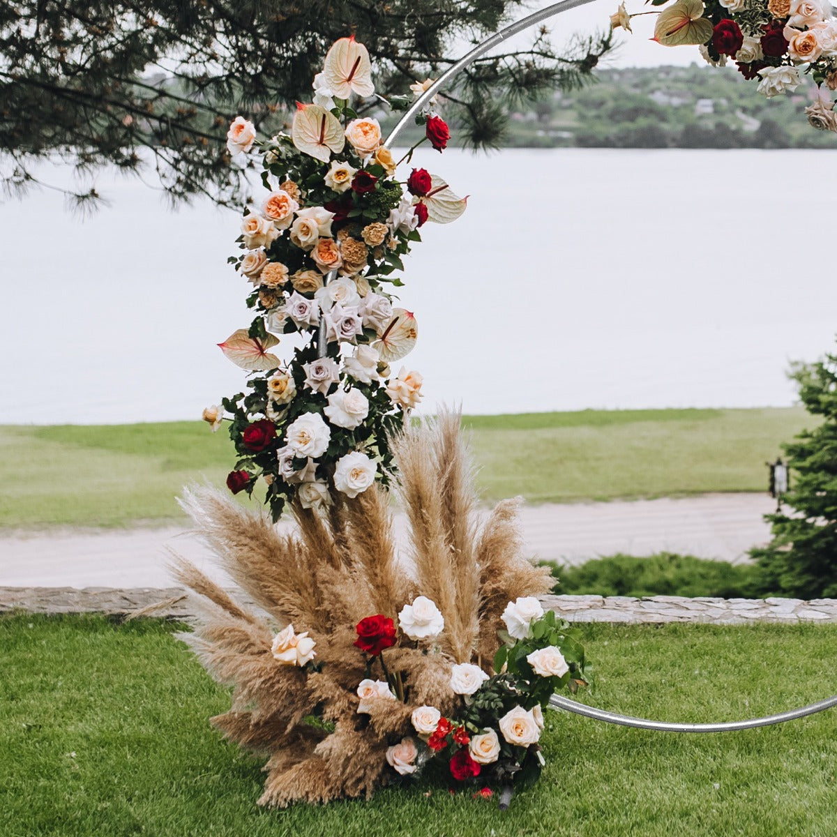 Eleganter Hochzeit Ballonbogen in Silber, geschmückt mit weißen, roten und rosa Blumen sowie getrocknetem Pampasgras, vor malerischem Wasserhintergrund