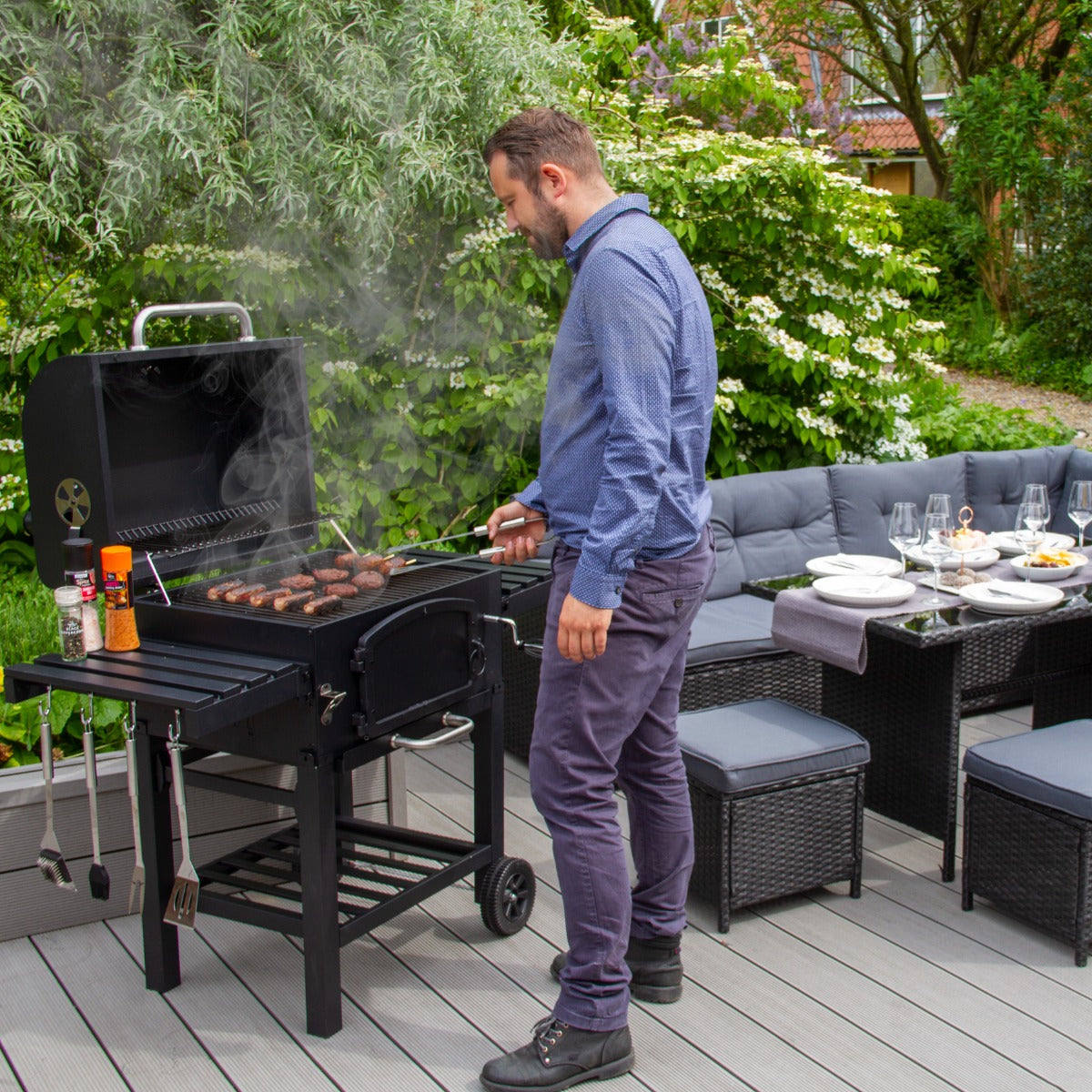 XL Holzkohlegrill Smoker in Aktion, mit einem Mann, der Würstchen grillt, umgeben von grünen Pflanzen und einer modernen Terrasse