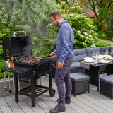XL Holzkohlegrill Smoker in Aktion, mit einem Mann, der Würstchen grillt, umgeben von grünen Pflanzen und einer modernen Terrasse
