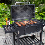 XXL Holzkohlegrill Smoker mit geöffnetem Deckel, rauchend, auf einem Holzdeck, mit gegrilltem Fleisch und Gewürzen daneben, umgeben von grüner Vegetation