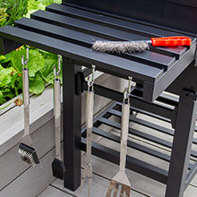 Schwarzer Grillwagen mit Edelstahl-Grillwerkzeugen, darunter Wender und Bürste, auf einer Holzterrasse umgeben von frischem Grün