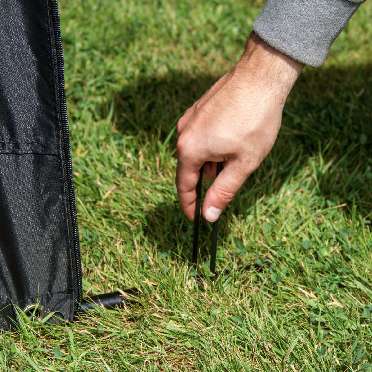 Ein Hand greift nach einem Zelthering, während er ein schwarzes Fahrradzelt auf grünem Gras aufstellt, um Stabilität zu gewährleisten