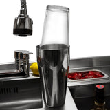 Elegante Cocktail-Bar Station mit glänzendem Edelstahl, transparentem Shaker und frischen Zutaten wie Zitronen und Erdbeeren im Hintergrund