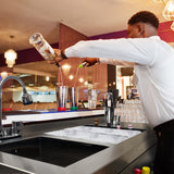 Elegante Cocktail-Bar Station mit einem Barkeeper, der Getränke in Edelstahl-Shaker gießt, umgeben von Gläsern und Eiswürfeln