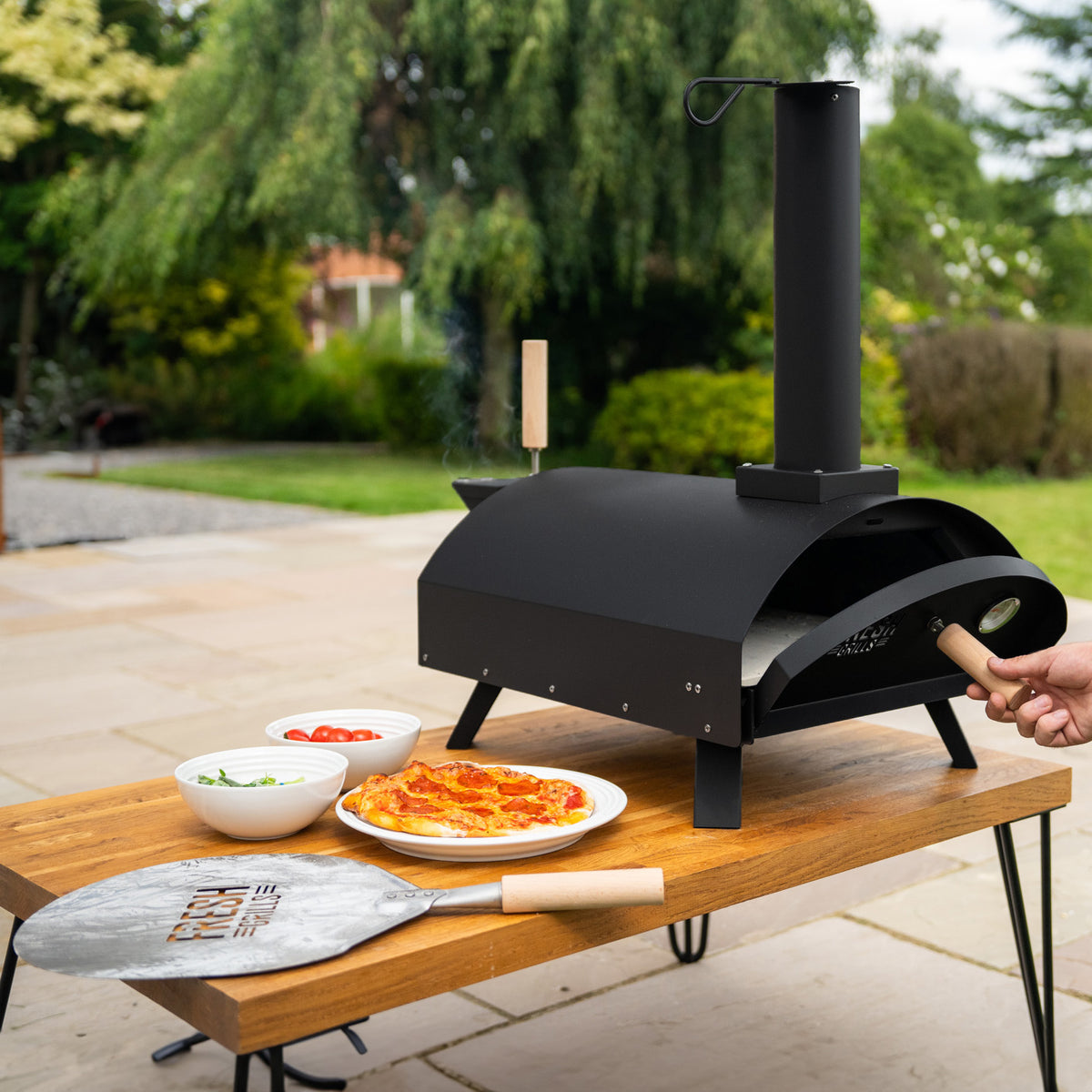 Klassischer Outdoor-Pizzaofen in mattschwarz, auf einem Holztisch, mit frischen Zutaten und einer Pizzaschaufel aus Edelstahl, ideal für Gartenpartys