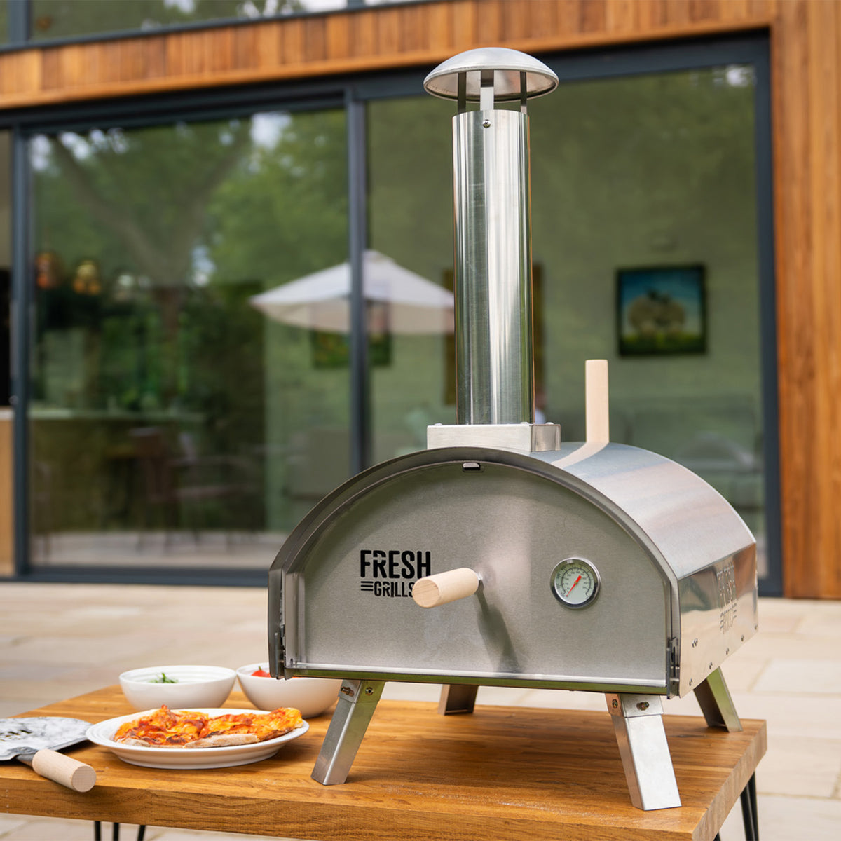 Eleganter Premium Outdoor-Pizzaofen aus rostfreiem Stahl mit einem hohen Schornstein, Thermometer und Holzgriffen, auf einem Holztisch mit Pizza und Saucen