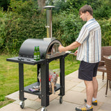 Freistehender Outdoor-Pizzaofen mit schwarzem Gehäuse, Vorbereitungsstation und einem Mann, der Pizza auf einem Holzofen backt, umgeben von Gartenmöbeln